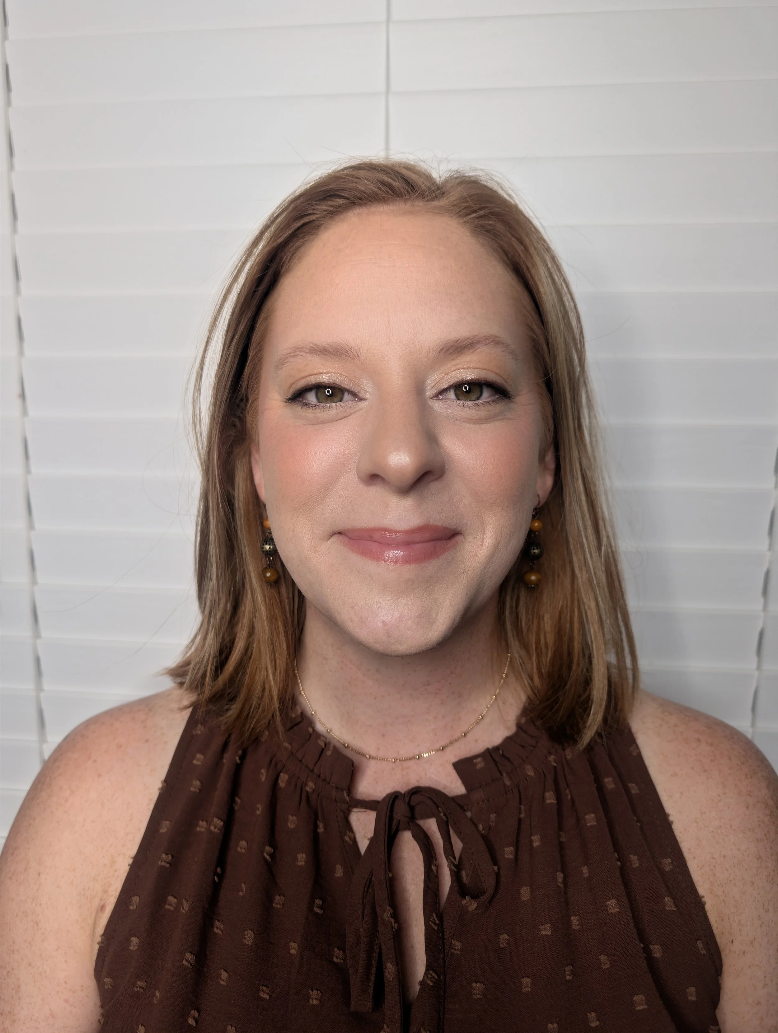 A woman with light brown hair, wearing earrings and a necklace, smiling at the camera in front of a white paneled background.