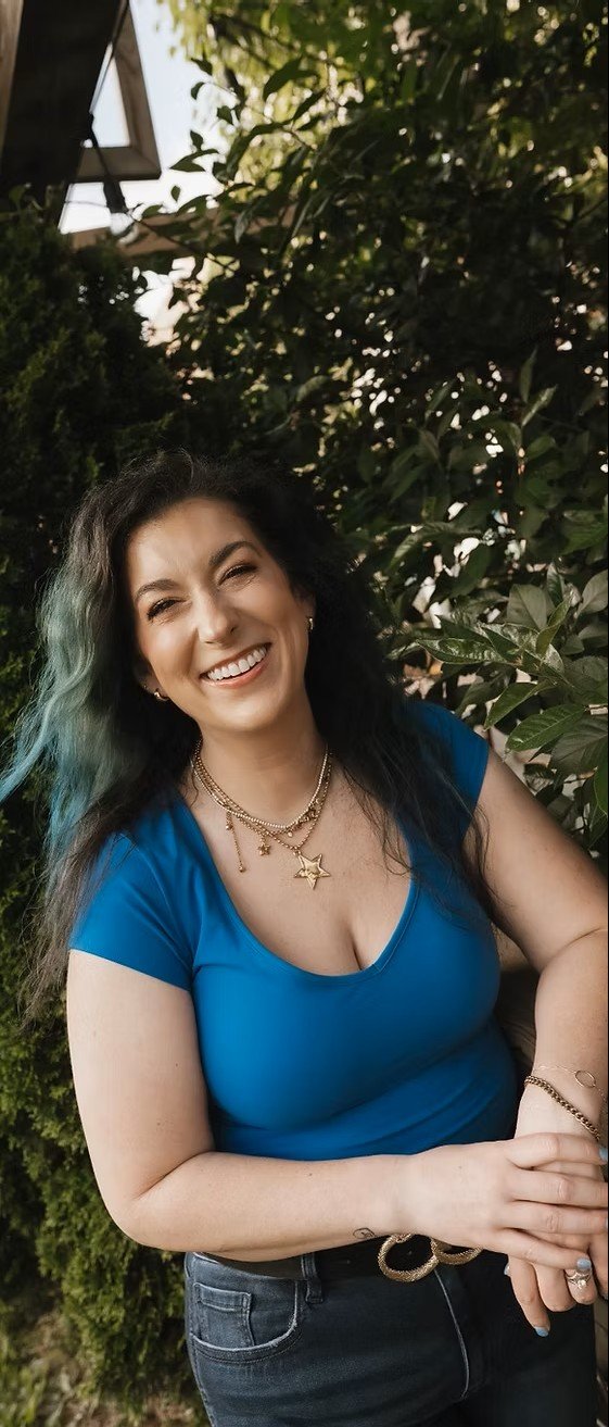 A woman with long dark hair and a blue top smiling outdoors, standing in front of green foliage and trees.