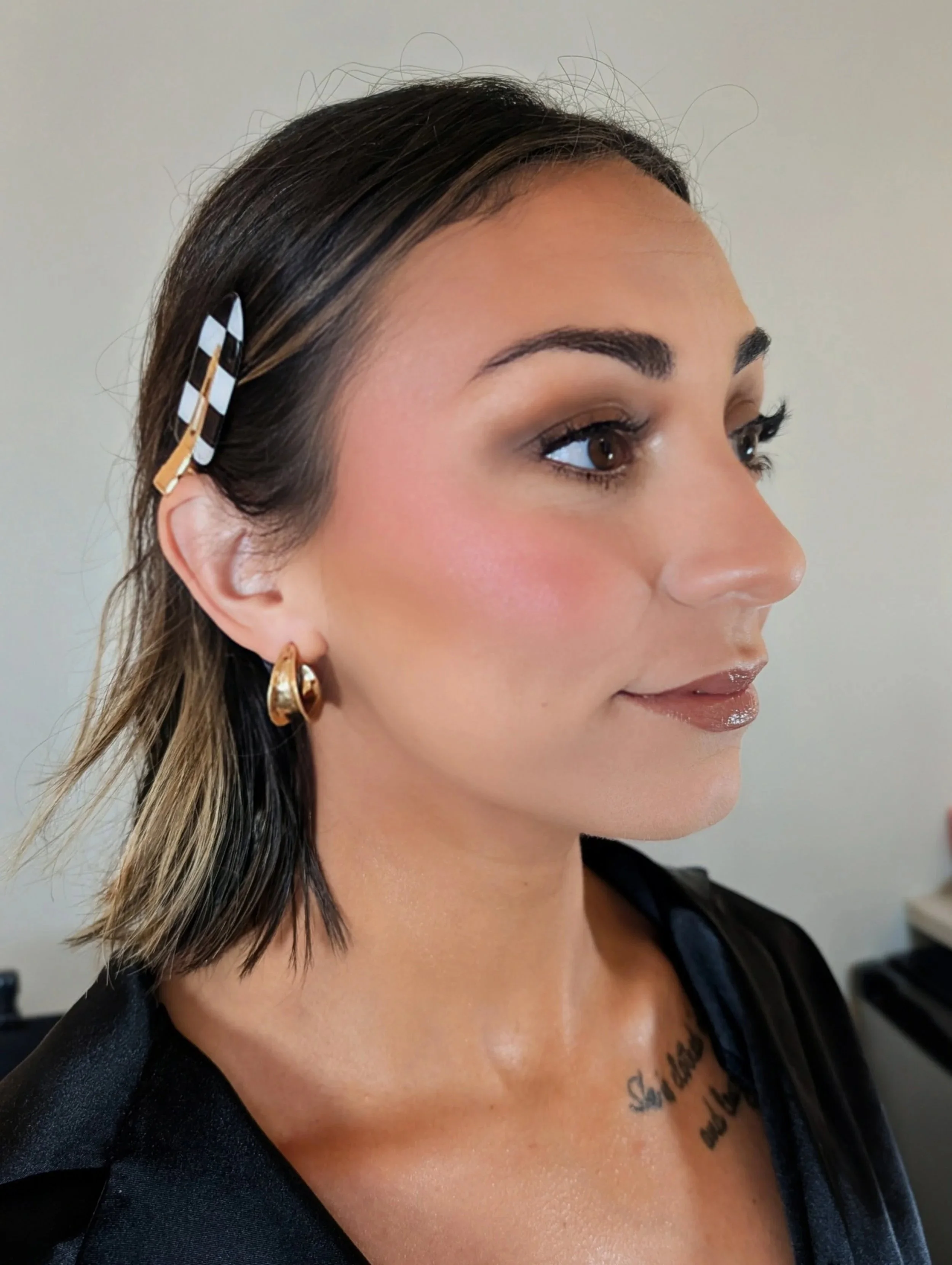 A woman with shoulder-length dark brown hair with highlights, secured on the side with a black and white checkered hair clip, wearing gold hoop earrings, a black top, and a tattoo partially visible on her chest, standing against a plain light-colored background.