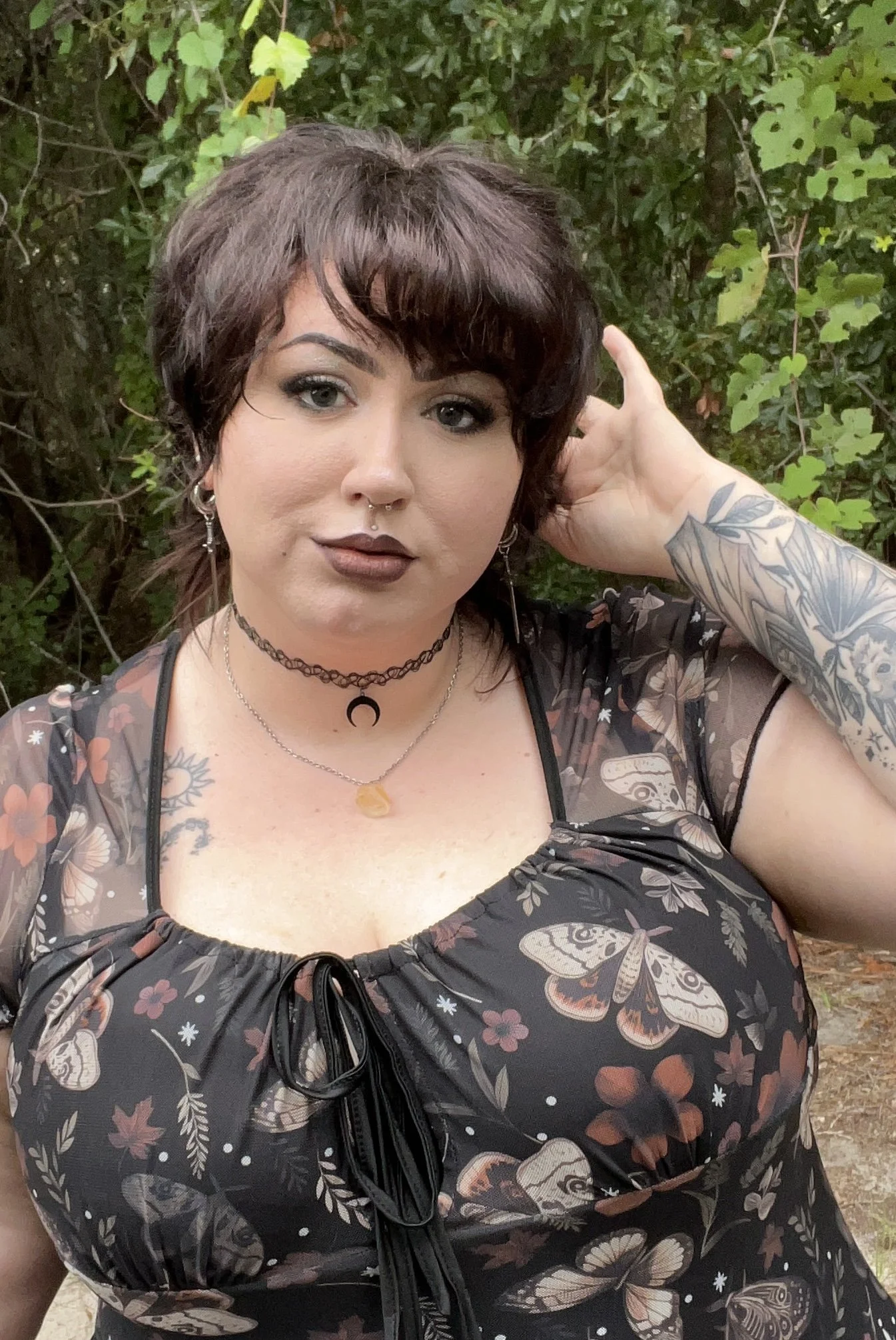 A woman with dark red, short, wavy hair, makeup, body tattoos on her arms and chest, and earrings, wearing dark floral clothing with a butterfly pattern, standing outdoors in front of green foliage.