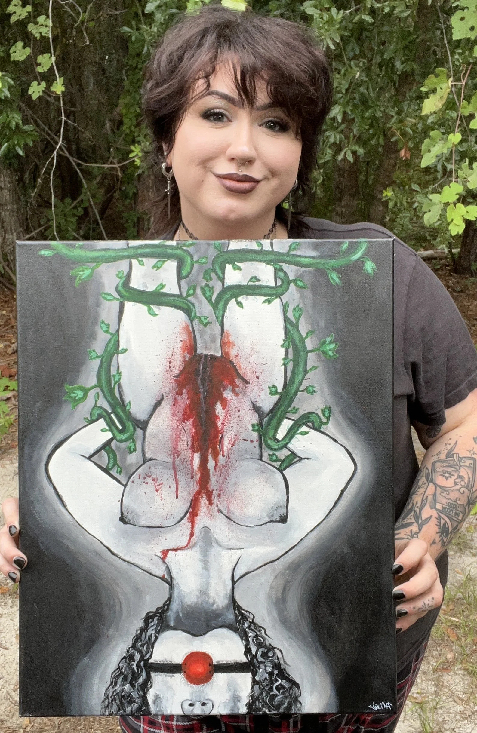 Woman with dark hair and makeup holding a painting of a detailed black and white female figure with green vines, blood, chains, and a red nose, outdoors with green foliage background.