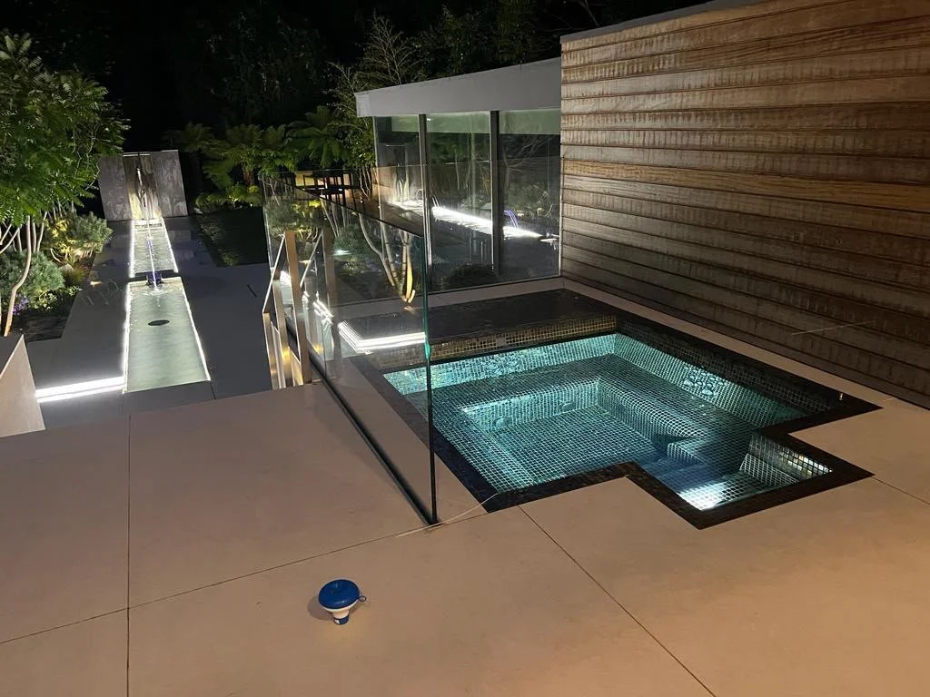 Night view of a modern outdoor pool area with a small hot tub, a waterfall feature, and landscaped greenery.