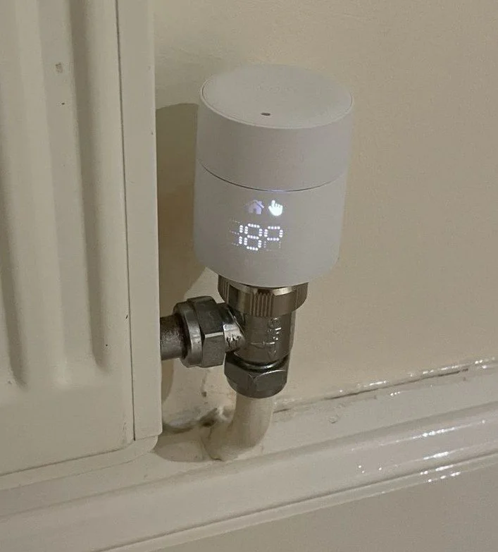 Smart thermostat attached to a heating radiator pipe, displaying temperature and control icons.