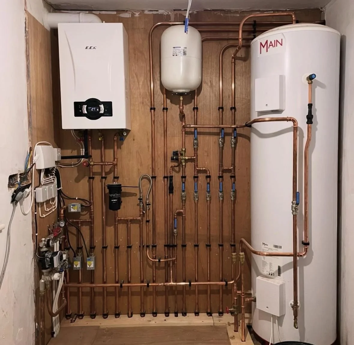 A plumbing and heating system with various copper pipes, boilers, and a water tank mounted on a wooden wall.