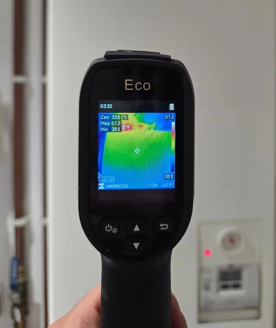 Infrared thermal imaging camera displaying temperature readings, with colors indicating heat distribution, held by a person in front of a white wall and electrical outlet.