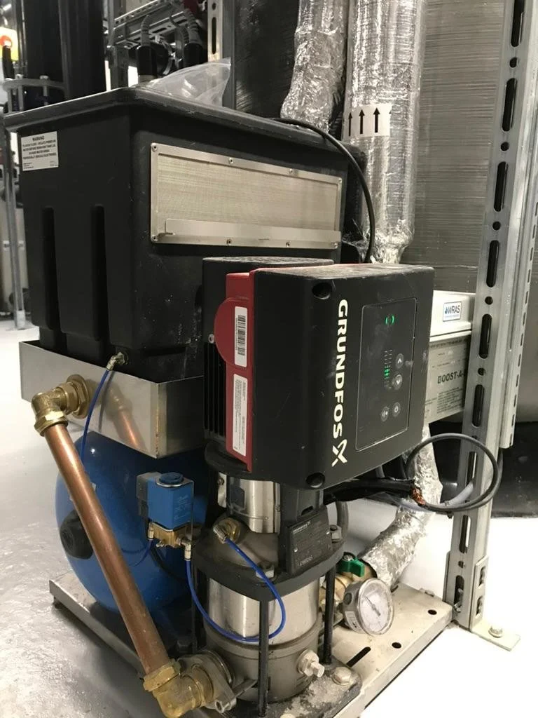 A piece of industrial equipment with a Grundfos pump, copper piping, and various gauges mounted on a metal frame in a facility with exposed ducts and shelving.