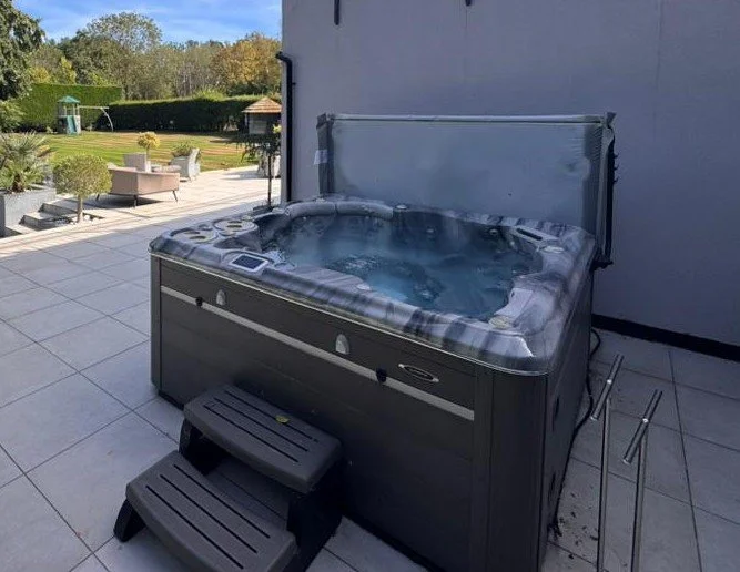 Outdoor hot tub with a cover, surrounded by patio tiles and set against a gray wall, with steps leading into the tub, and a garden with trees and outdoor seating in the background.