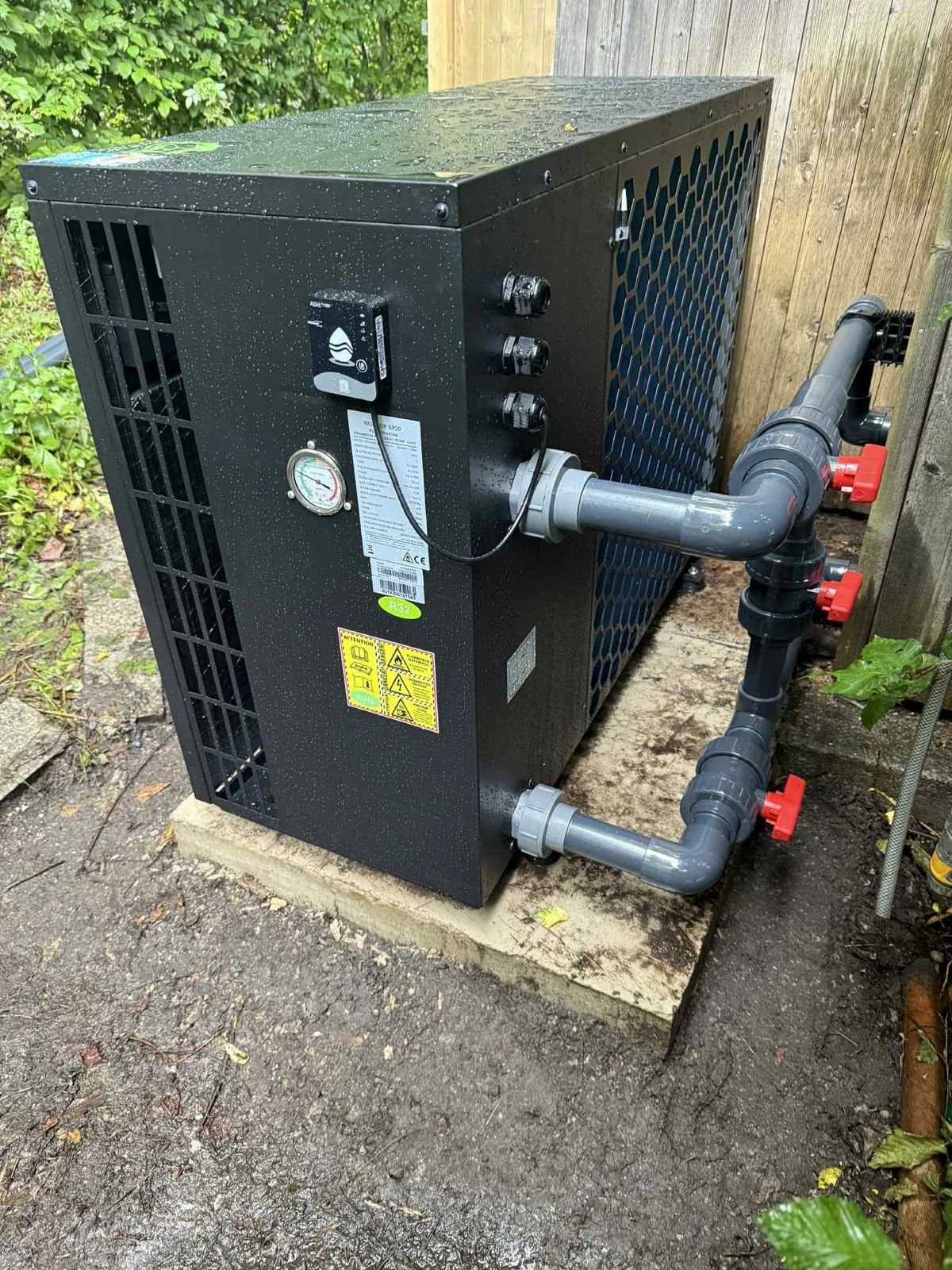 An outdoor pool heat pump unit installed on a concrete pad, with black piping and red valves connected, surrounded by a wooden fence and green foliage.