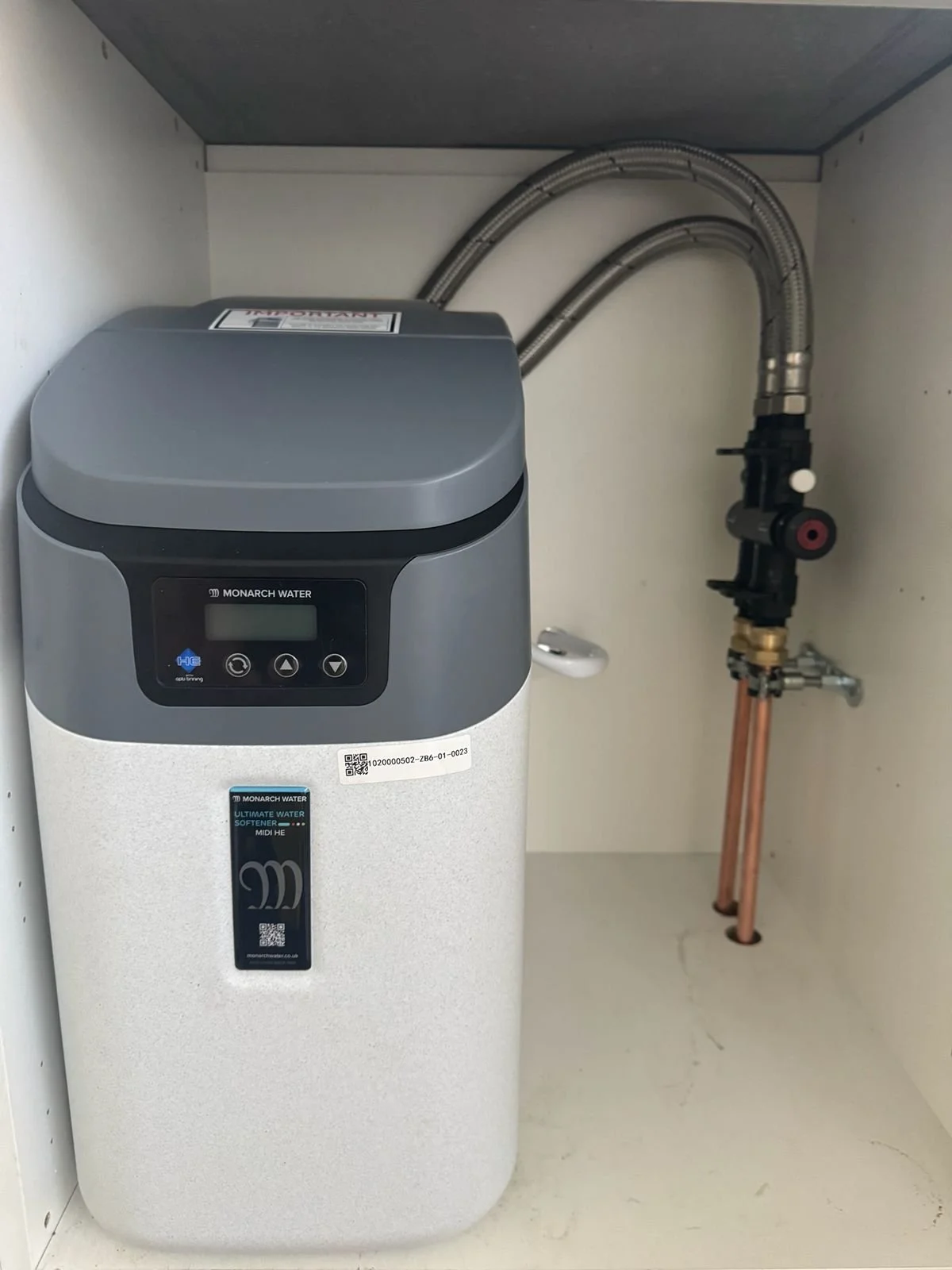 An under-sink water softener system with copper pipes and flexible hoses connected to plumbing. The device has a digital display and controls.