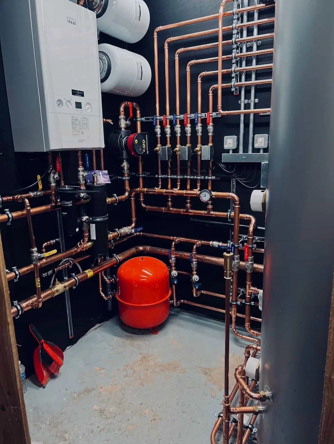 A plumbing or heating system with copper pipes, gauges, valves, a red expansion tank, and a white boiler unit installed on a dark wall.