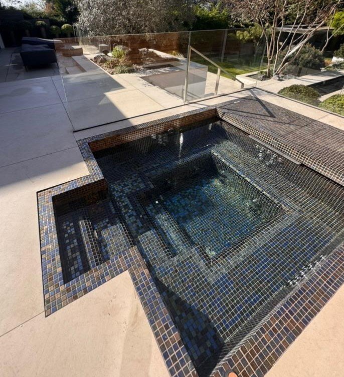 Empty outdoor hot tub with mosaic tiles, surrounded by a concrete patio, glass railing, and garden with trees and shrubs.