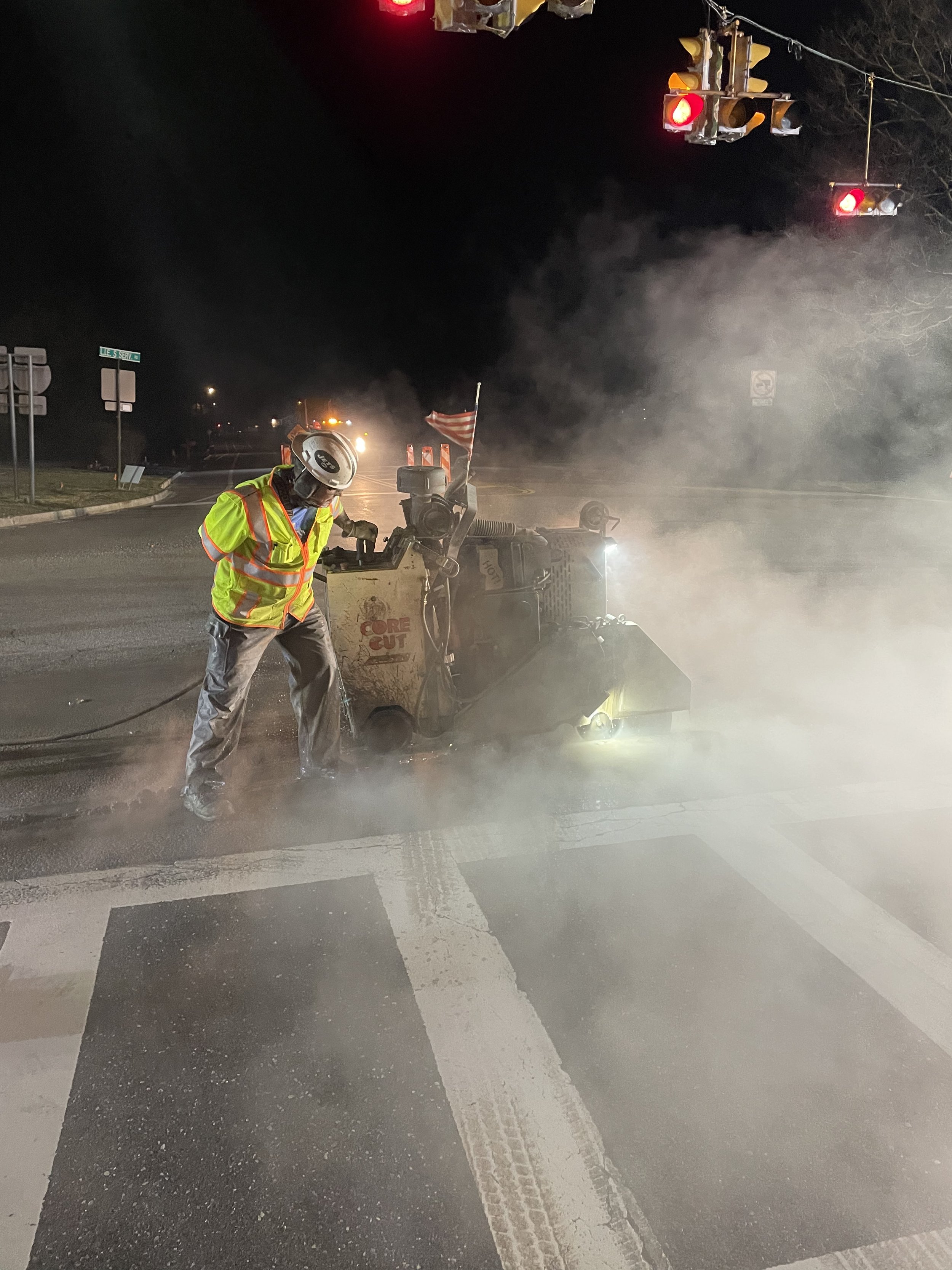 A worker in a yellow safety vest and helmet operating a street line cleaning machine at night. Smoke and dust are visible, and traffic lights are red. The scene takes place at an intersection, with street signs and road markings visible.