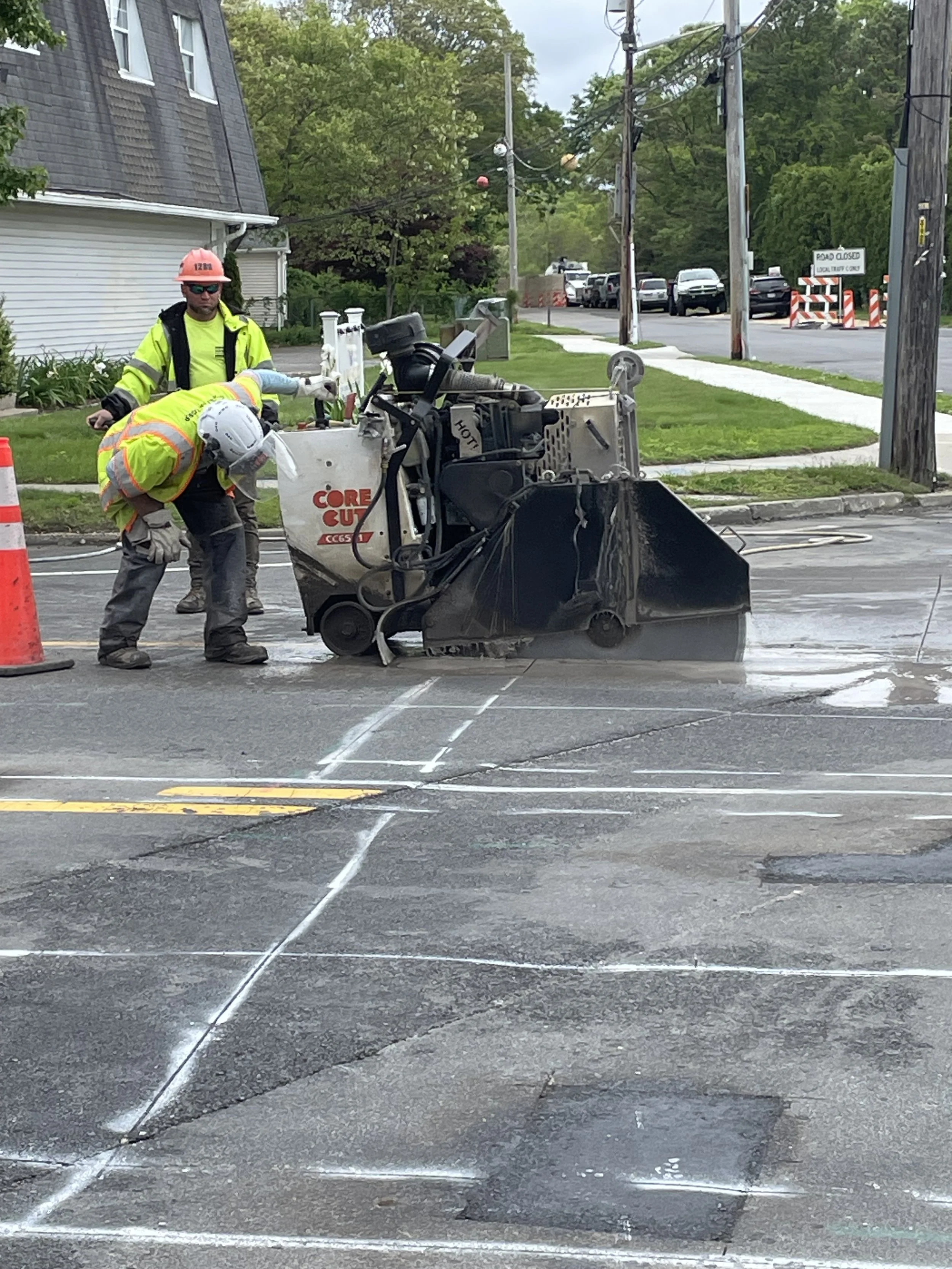 Two construction workers are working on a road, using a large street sweeper machine. One worker is bending down, wearing a white helmet and fluorescent vest, while the other stands behind, wearing a pink helmet, glasses, and a yellow safety jacket. There are orange traffic cones and a lined chalk or paint marking the road surface. In the background, there are trees, parked cars, utility poles, and a building.