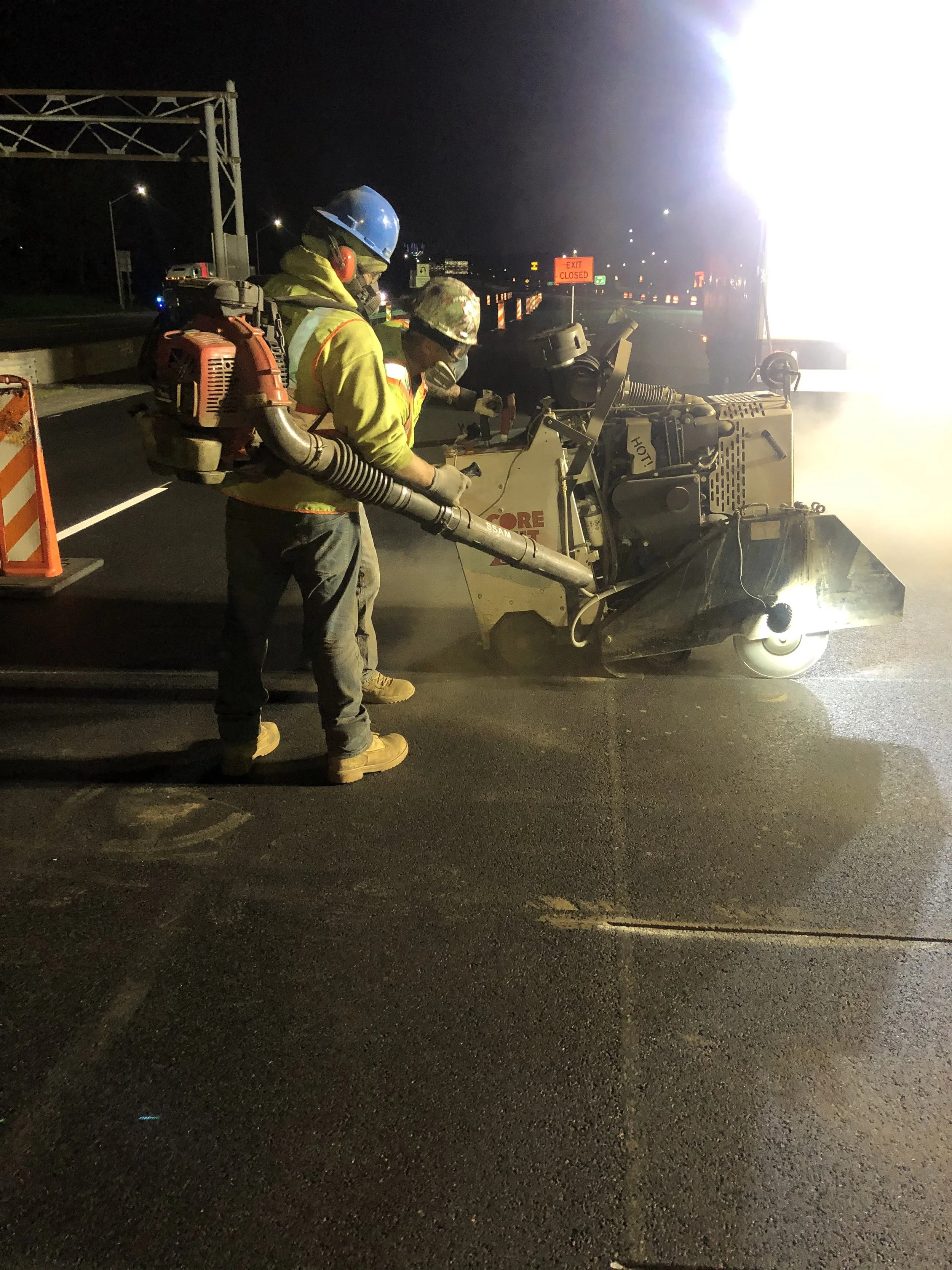 Two construction workers in yellow safety jackets and helmets, working on a road at night with bright lights, orange barricades, and an 'Exit Closed' sign in the background. One worker is operating a road milling machine.