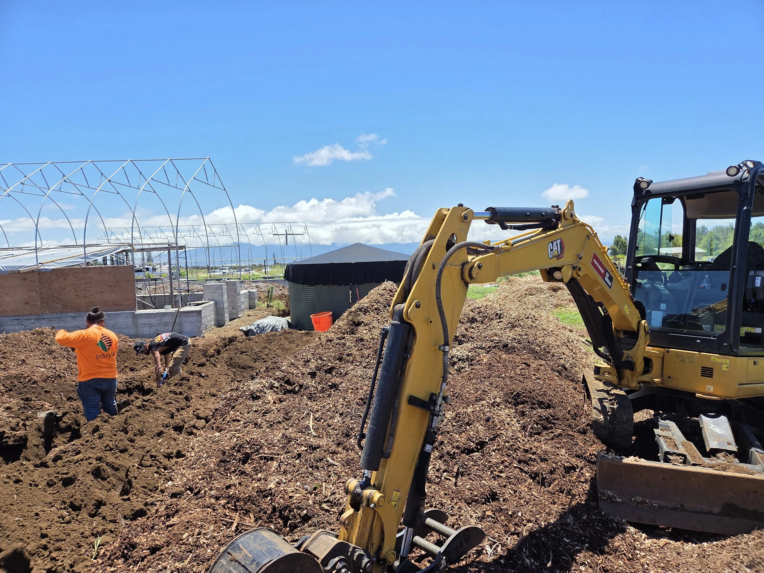 Construction site with a yellow excavator, two workers digging in the dirt, greenhouses in the background, blue sky with some clouds.