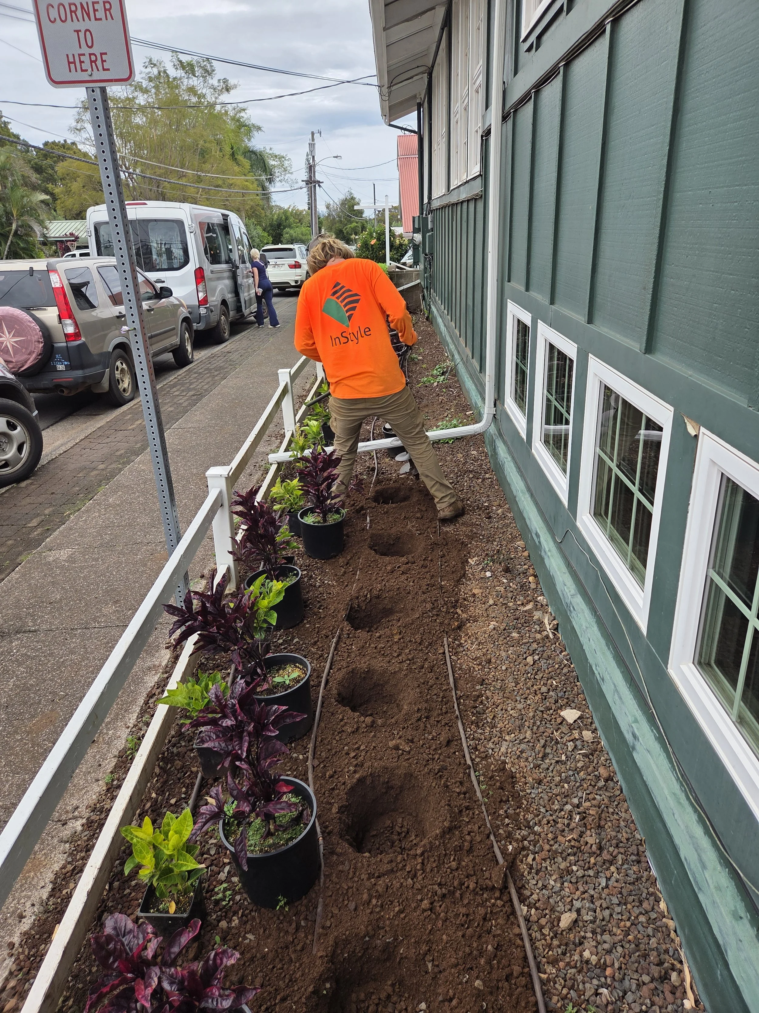 A person planting small flowering plants along a sidewalk next to a green building with multiple windows. The person is wearing an orange jacket with a logo on the back that says 'InStyle.' Several planted pots are arranged in a row, and the person a