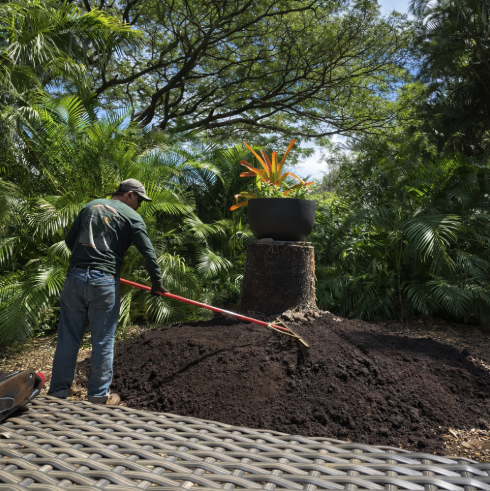 Man gardening and planting soil around a potted plant in a lush, green outdoor setting.