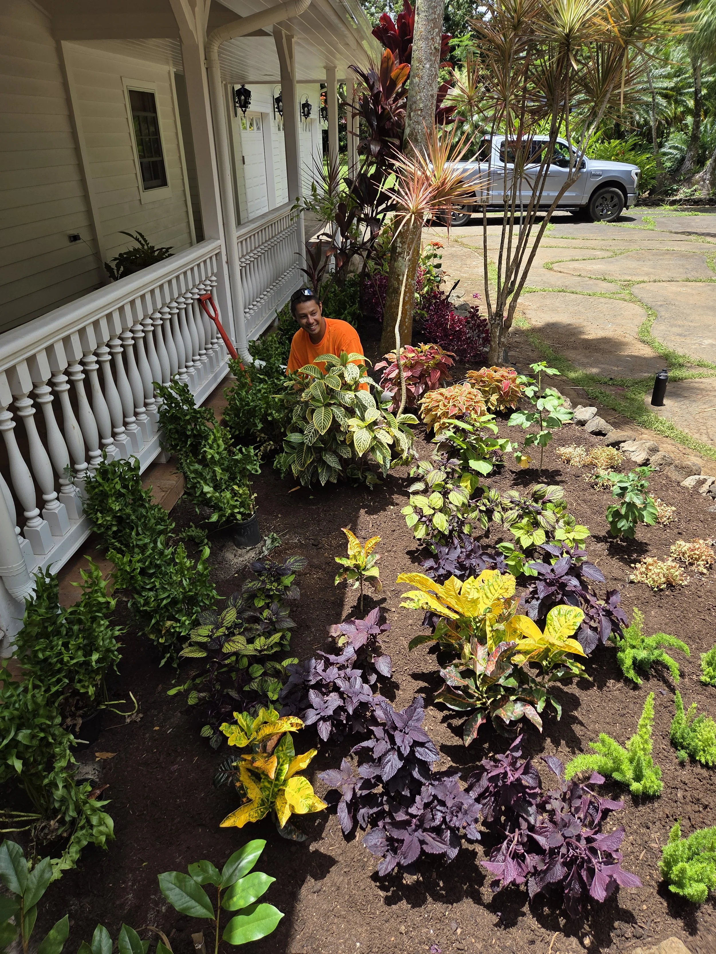 A person wearing an orange shirt is gardening in a lush backyard with a variety of colorful plants and flowers. There is a white house with a porch and a parked silver truck in the background.