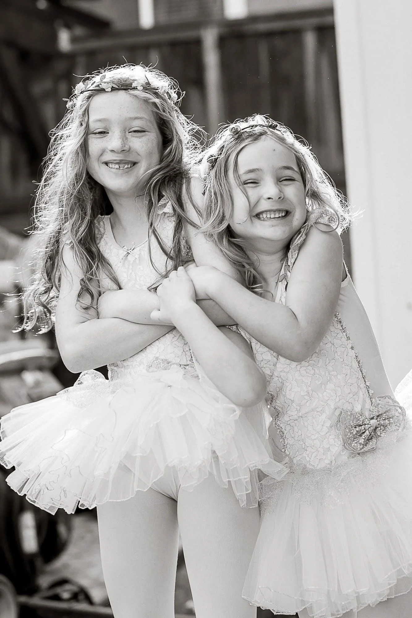 Two young girls in ballet tutus and floral crowns smiling and hugging each other in a black and white photo.