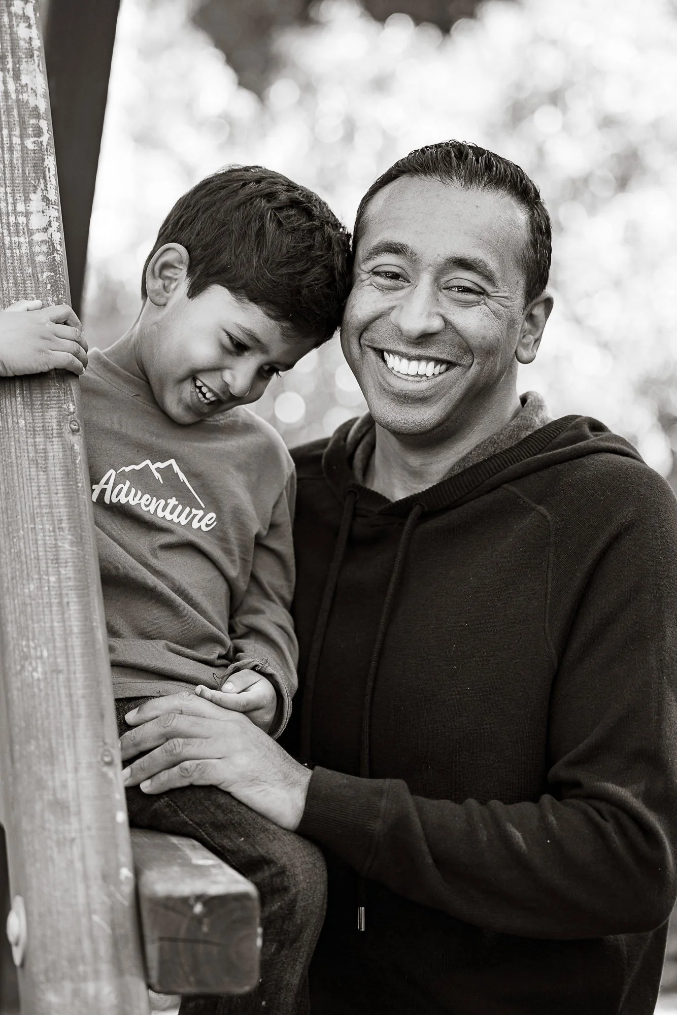 A happy man and a smiling boy sharing a joyful moment outdoors, with the boy leaning against a wooden railing and the man's hand gently touching the boy's knee.