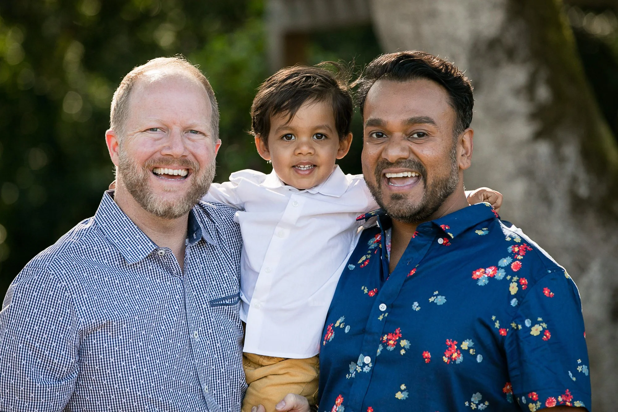 Three men and a young child smiling outdoors with green foliage in the background.