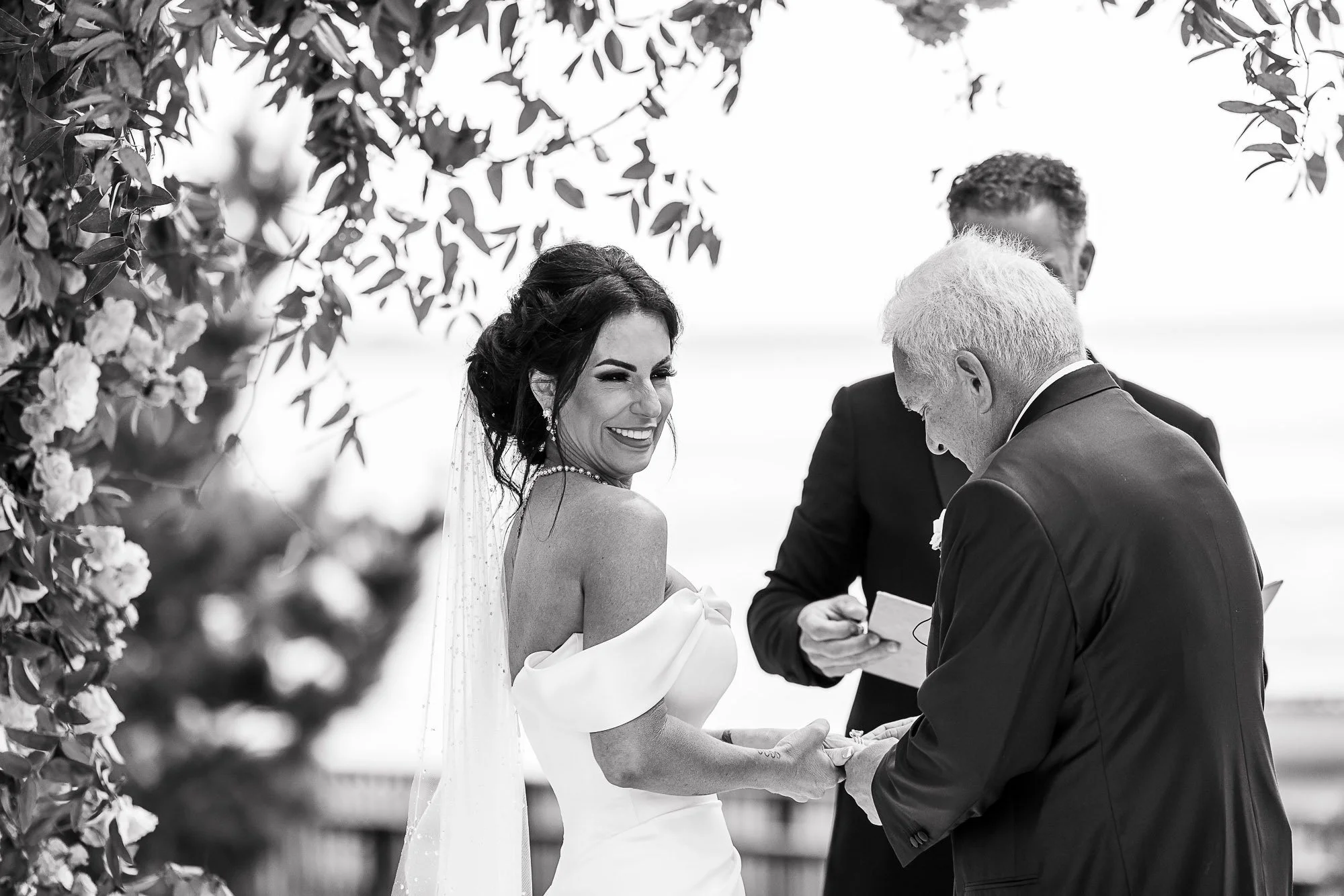 A wedding ceremony outdoors with a bride and groom exchanging vows. The bride is smiling and holding hands with the groom, who is reading from a paper. There are guests and foliage visible in the background, all depicted in black and white.