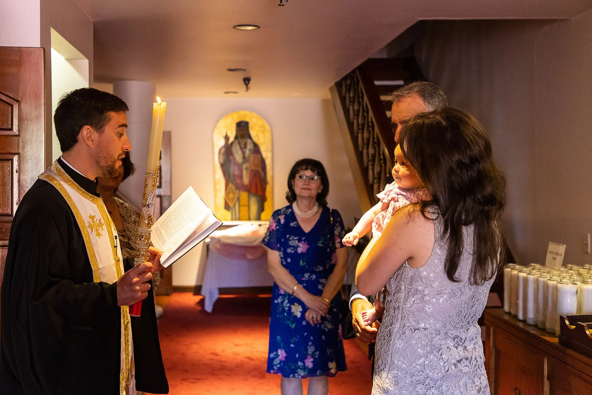 A religious ceremony inside a church with a young man dressed in religious attire reading from a book, a woman holding a smiling young girl, and two women in the background observing the event.