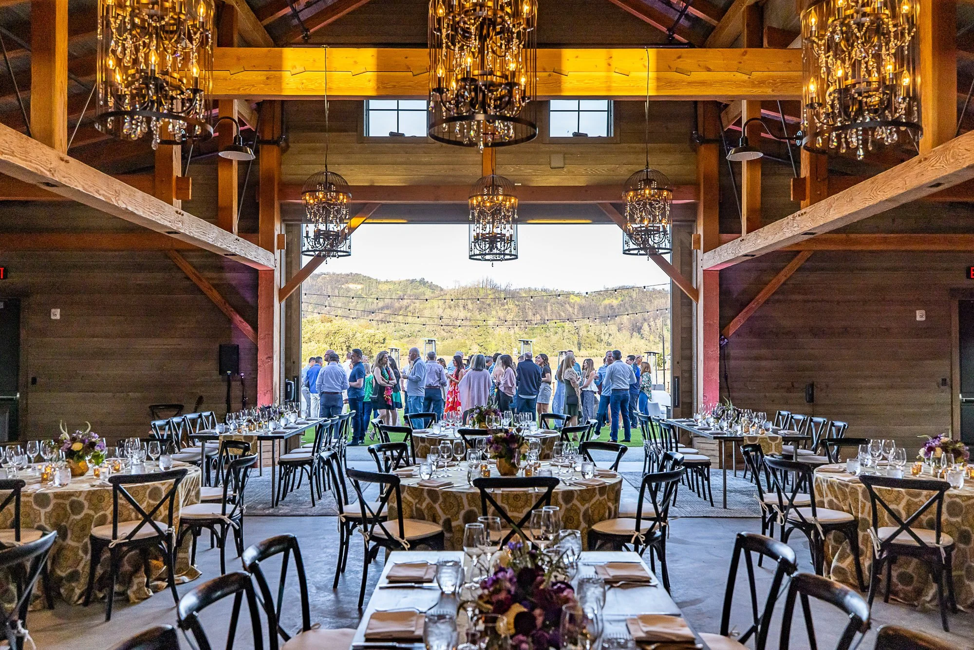 Indoor event space at the Napa Valley Four Seasons with round tables set for a celebration, decorated with flowers and glassware, with a large open doorway leading outside where a group of people are gathered chandeliers overhead.