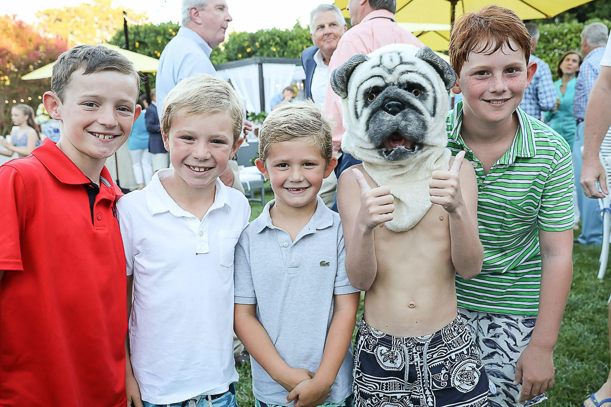 A group of young boys standing together, smiling at the camera, at an outdoor event during the daytime. One boy is shirtless and wearing a dog costume headpiece resembling a bulldog, giving two thumbs up. The background shows other people, trees, and