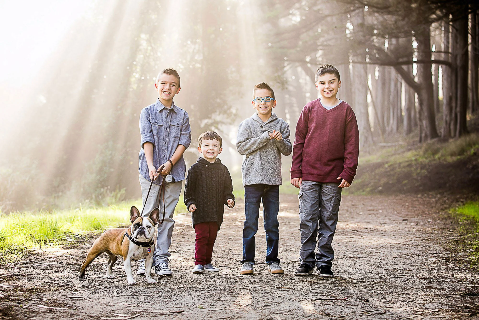 Five children walking on a dirt trail in a forest with a bulldog. The children are smiling and dressed casually, with sunlight filtering through trees in the background at Marine Fitzgerald Preserve in Half Moon Bay.