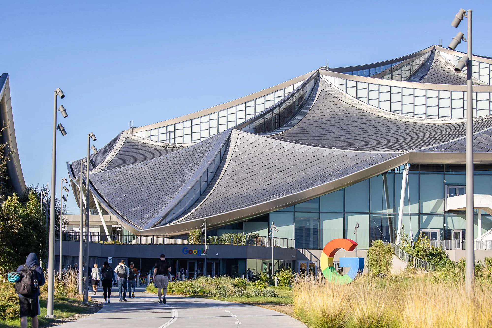 Google headquarters building with distinctive modern architecture featuring a curved, metal roof and glass windows, surrounded by people walking on a pathway and greenery, with the Google logo visible.
