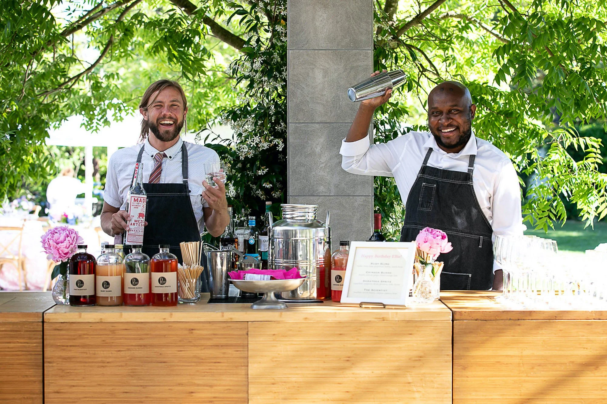 Two men are standing behind a wooden bar counter outdoors, smiling and wearing aprons. The man on the left has long hair and a beard, holding a clear glass, while the man on the right is bald, using a cocktail shaker. The bar has bottles of drink, fl