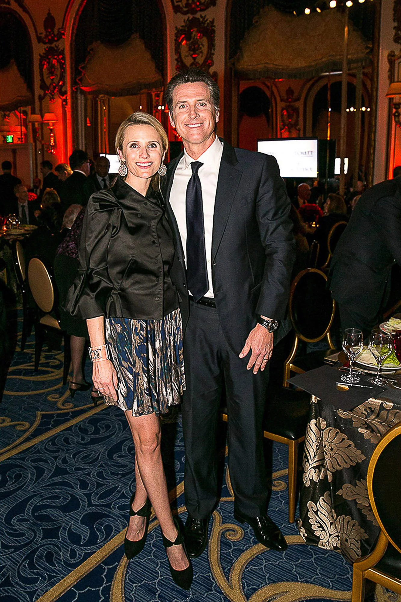 Gavin Newsom and wife together at a formal event in a decorated ballroom with ornate gold and dark wood accents. The woman is wearing a black satin jacket over a colorful skirt, and the man is in a dark suit with a white shirt and a black tie. 
