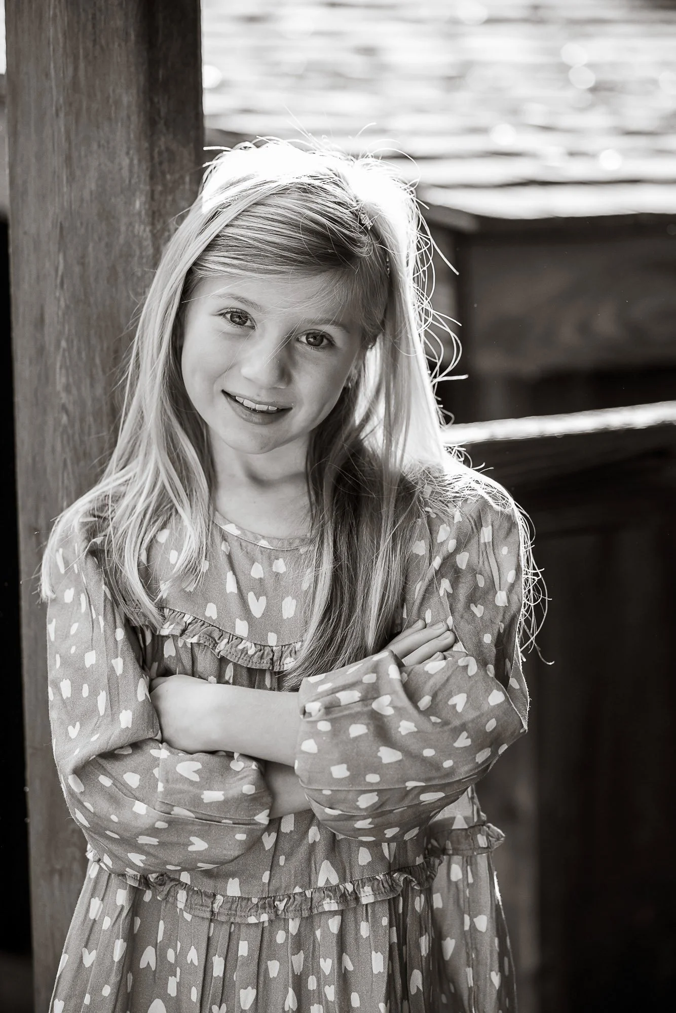 A black and white photo of a young girl with long, light-colored hair, smiling and crossing her arms, standing outdoors near a wooden post with water in the background.