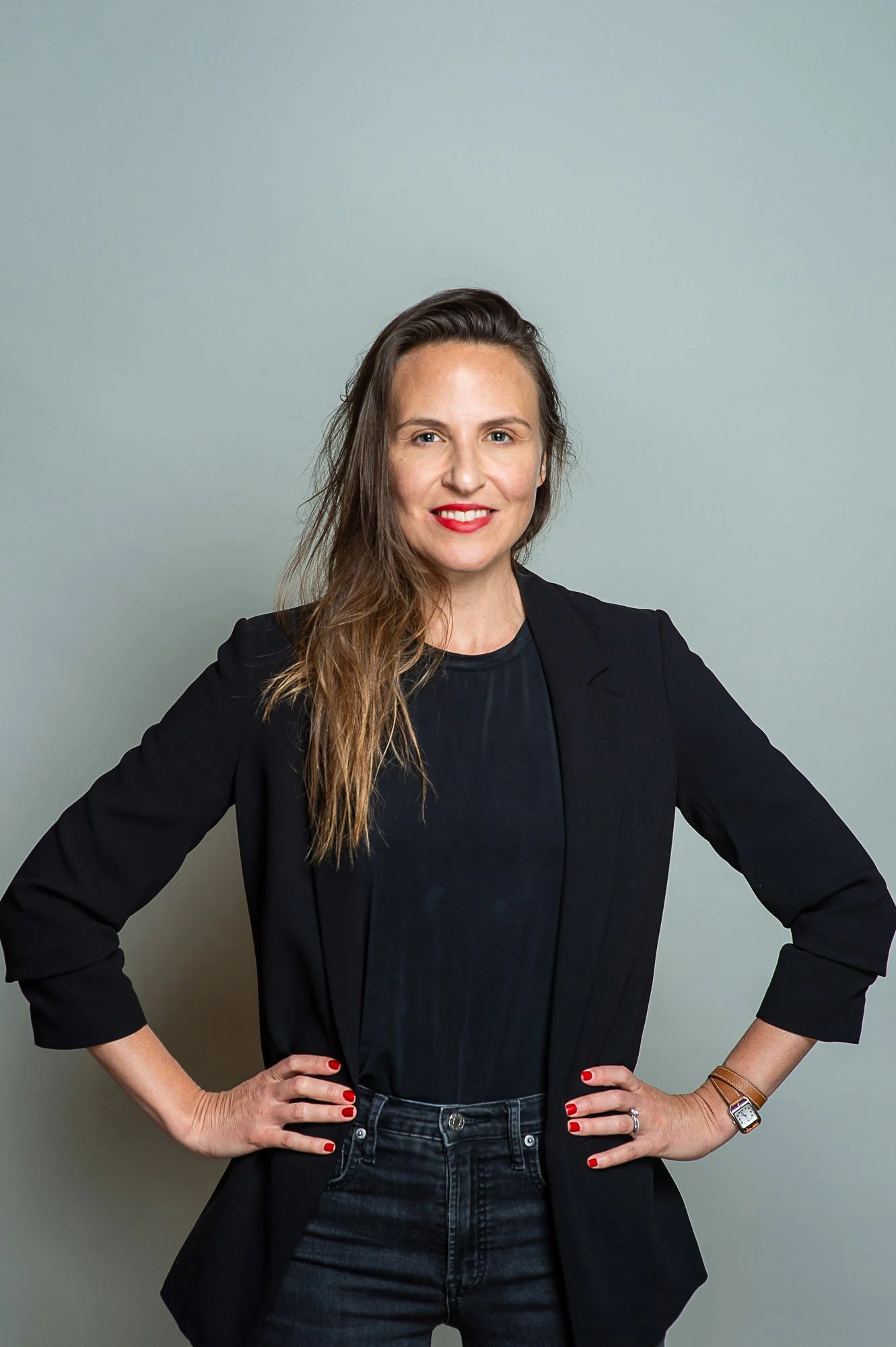 A woman standing with her hands on her hips, wearing a black blazer and jeans, smiling with red lipstick and red nail polish, against a plain light grey background.