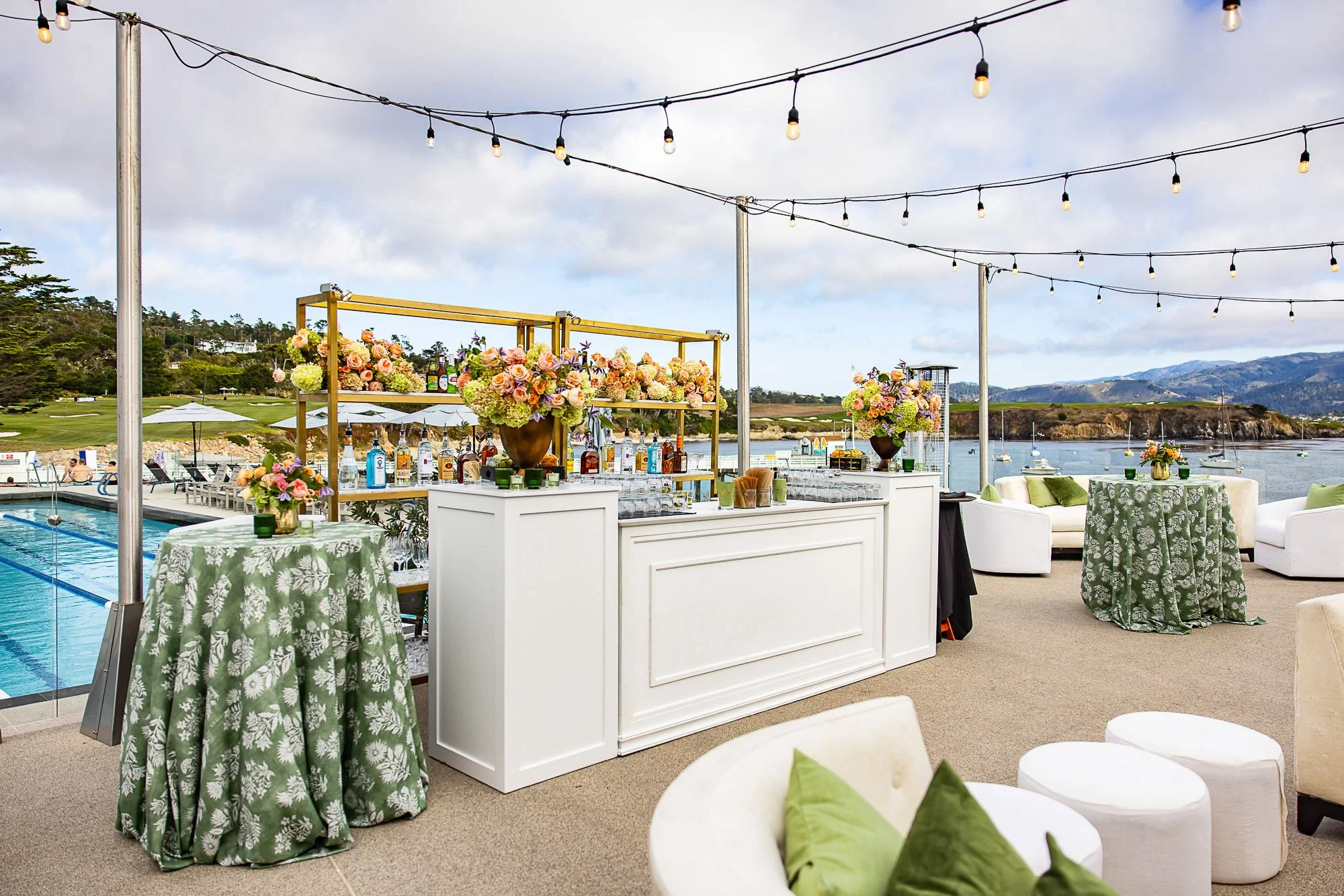 Outdoor waterfront party area with string lights, floral arrangement bar, and seating with green and white decor, overlooking mountains and water at the Beach and Tennis Club in Pebble Beach, CA.