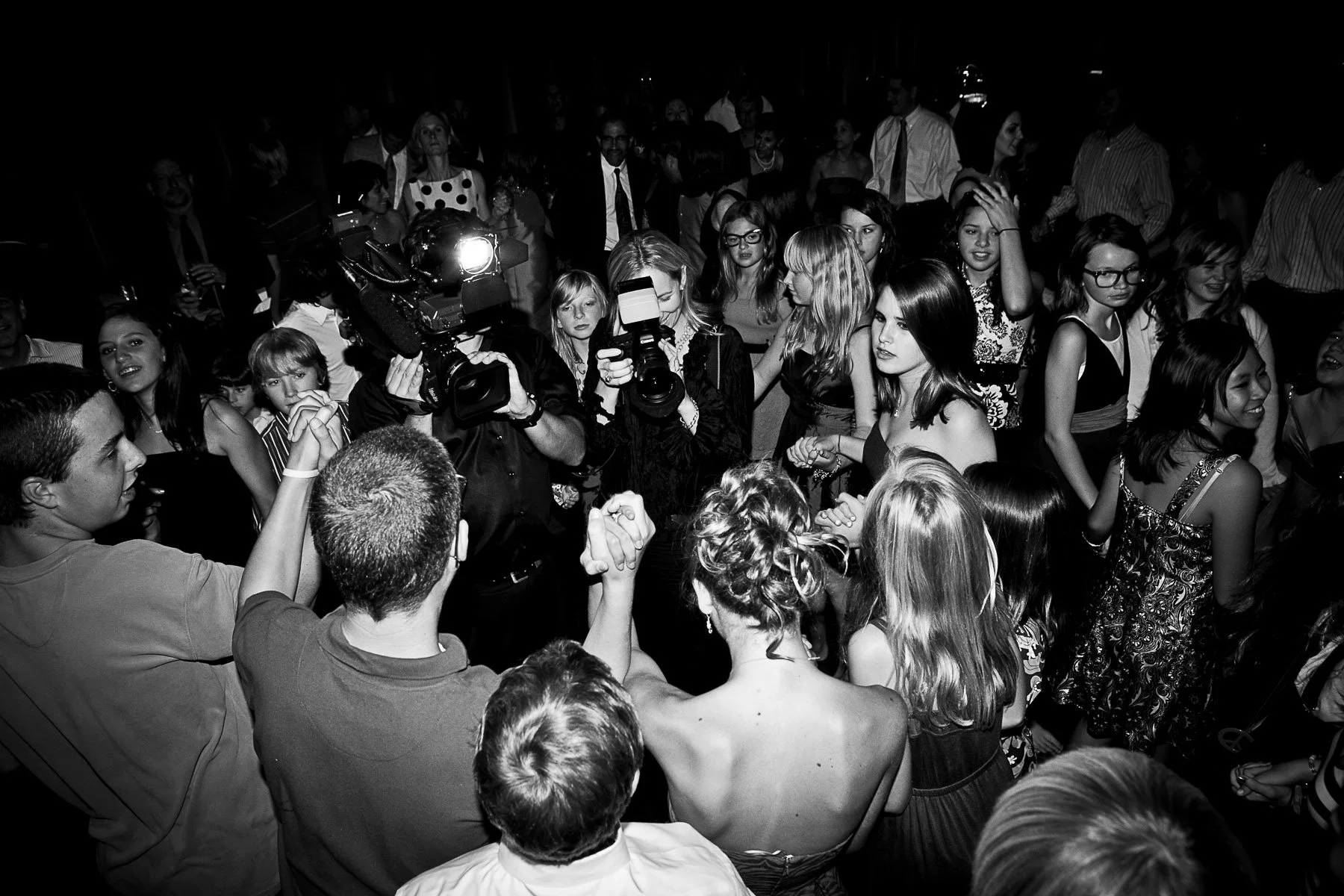 Tara and Martin photographing and recording video the guests dancing with arms raised at a mitzvah reception, black and white