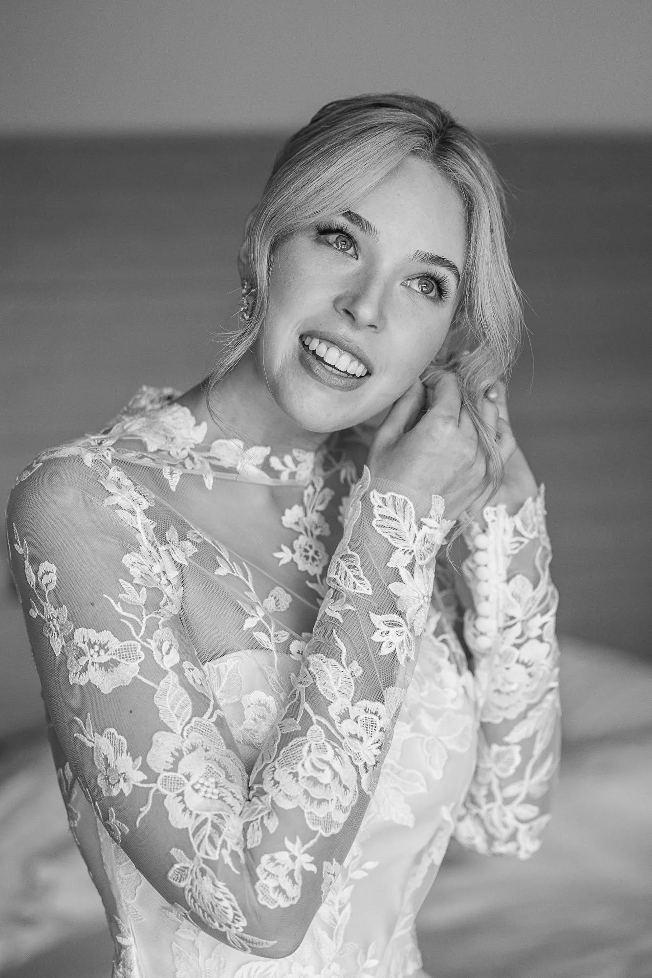 A woman with short blonde hair and bright eyes smiling. She is wearing a lace dress with floral embroidery and is touching her hair near her ear at Cavallo Point in Sausalito, CA.