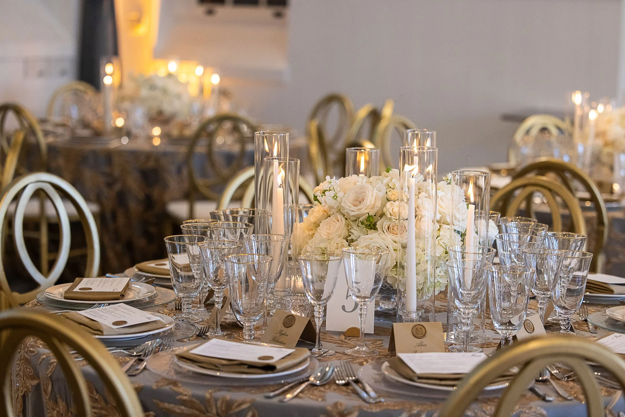 A banquet table set for a formal event with glassware, plates, and napkins, featuring a centerpiece of white flowers and tall candles, surrounded by gold chairs, in a decorated event space at Cavallo Point in Sausalito, CA.
