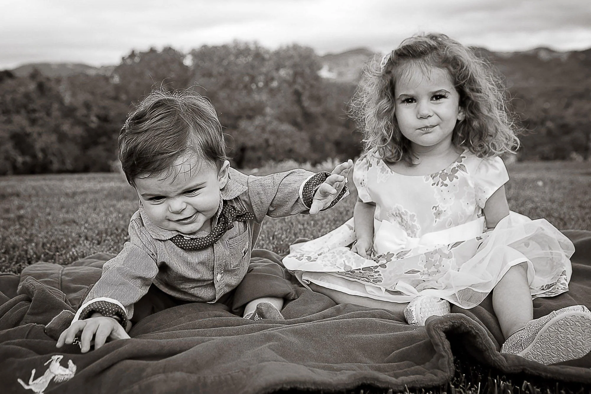 Two young children, a boy and a girl, sitting outdoors on a blanket in a grassy area with trees and mountains in the background. The boy is reaching out with his hand, and the girl is sitting beside him, both looking at the camera. The image is in bl