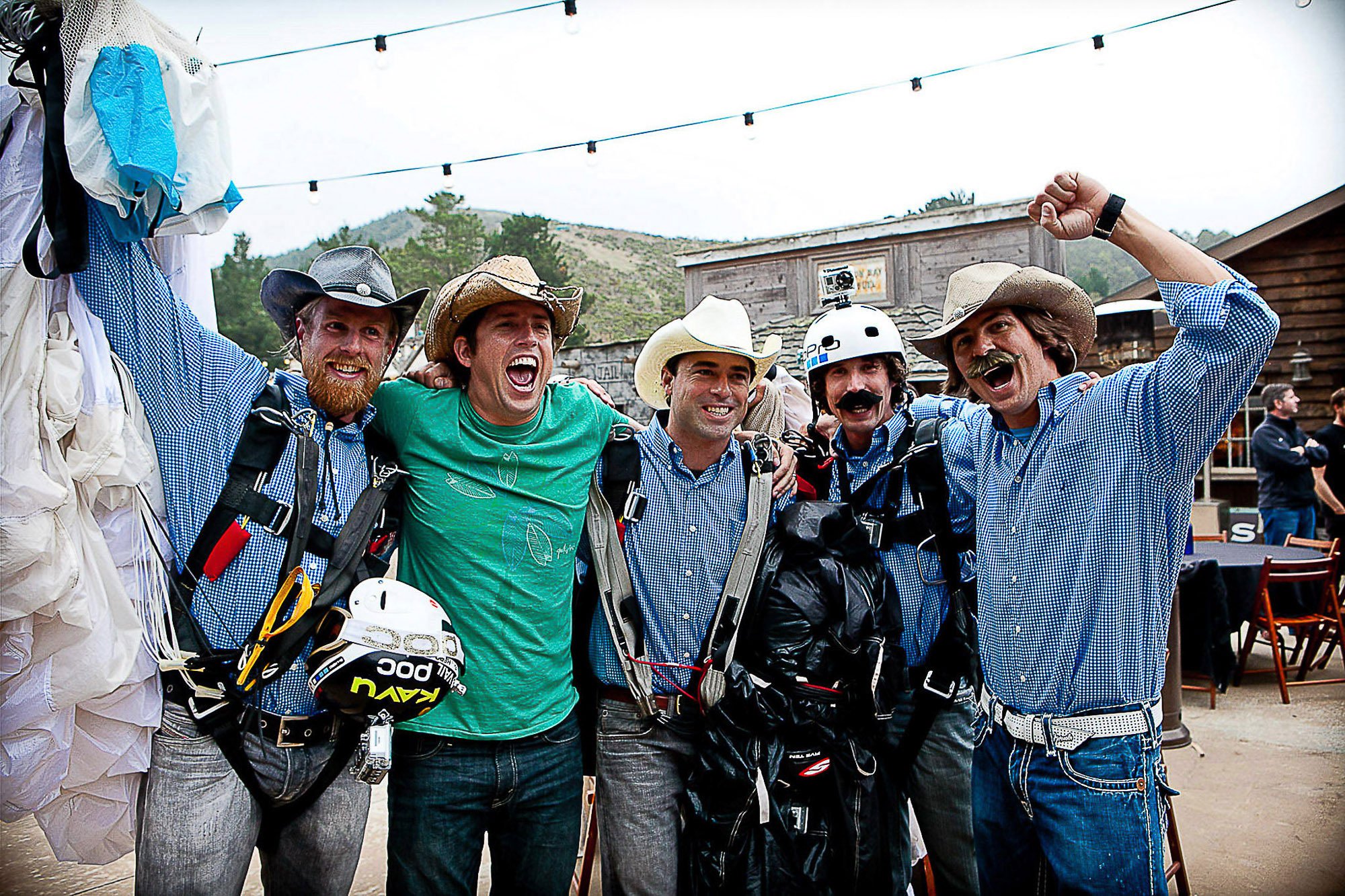 Nick Woodman and a group of four men from GoPro wearing cowboy hats and casual clothes, smiling and standing close together outdoors, with mountains and wooden buildings in the background. Some are wearing safety harnesses and helmets.