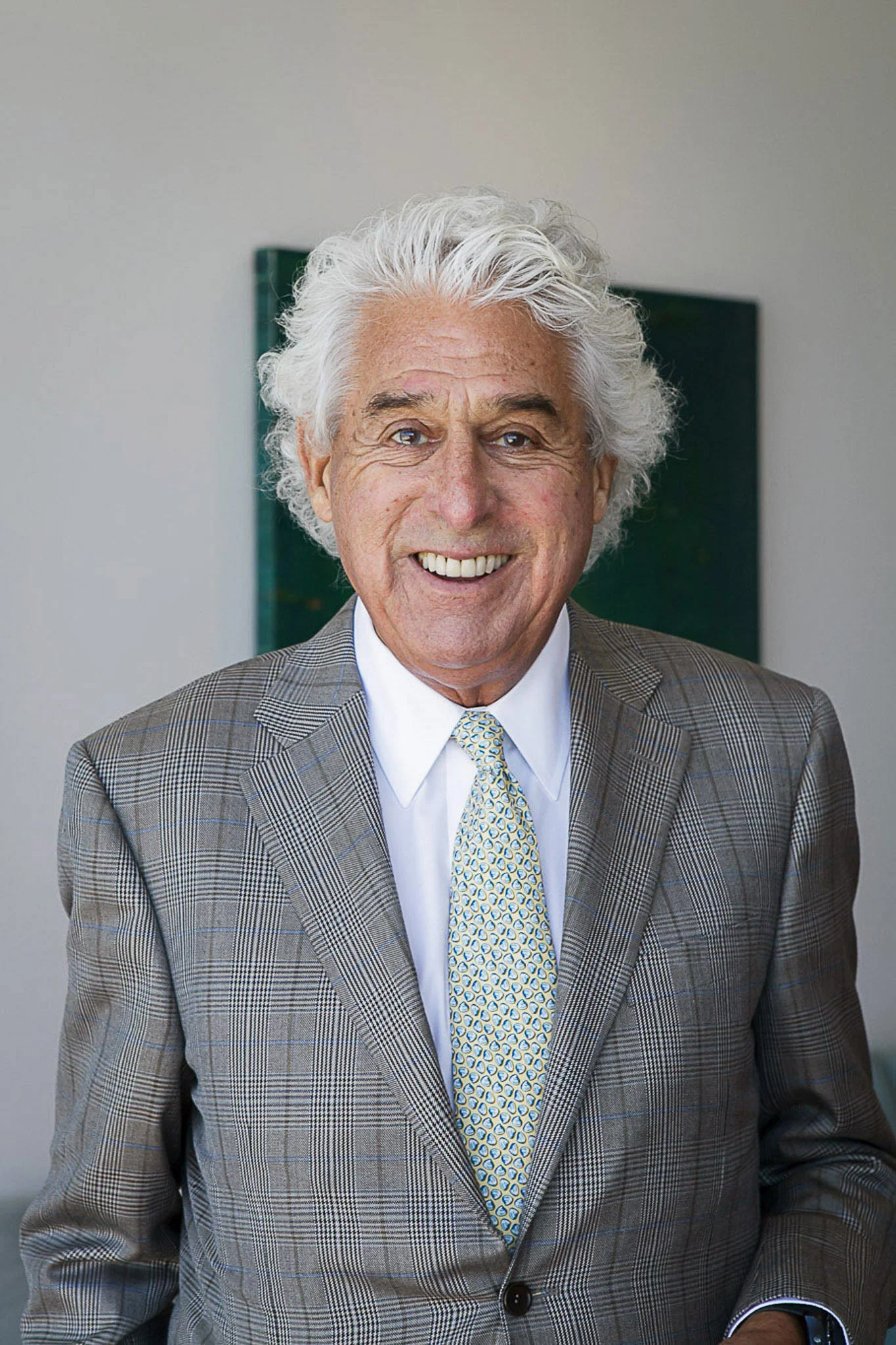 A corporate headshot of a smiling elderly man with white curly hair wearing a grey plaid suit, white shirt, blue patterned tie, and a watch on his left wrist.