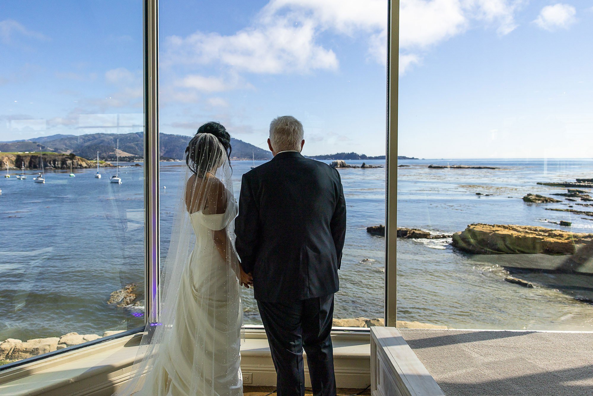 A bride and groom standing hand in hand, facing a large window with a view of water and boats, during their wedding ceremony at the Beach and Tennis Club in Pebble Beach, CA.