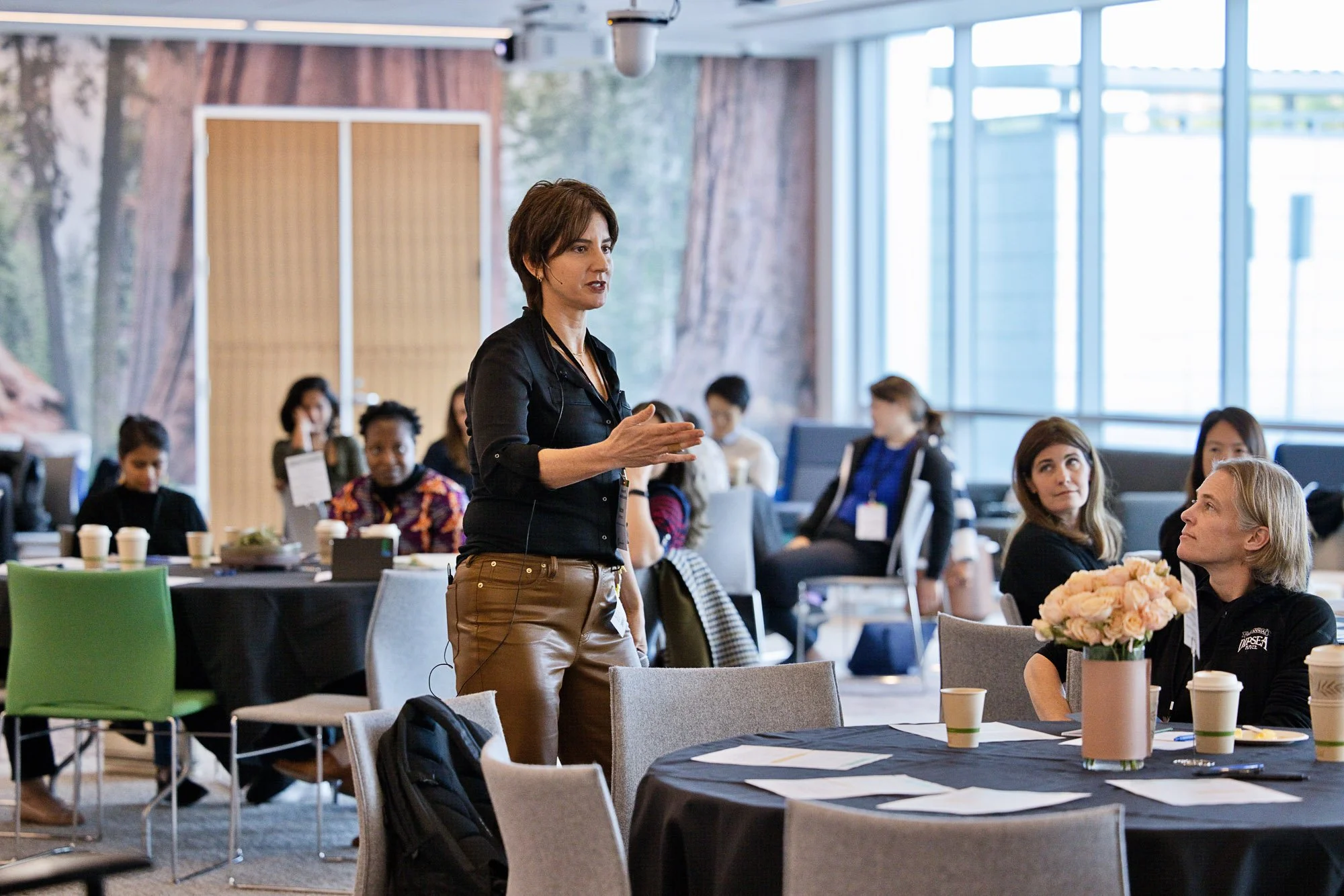 A woman in a black shirt and brown pants speaking to an audience in a conference room with large windows and attendees seated at round tables.