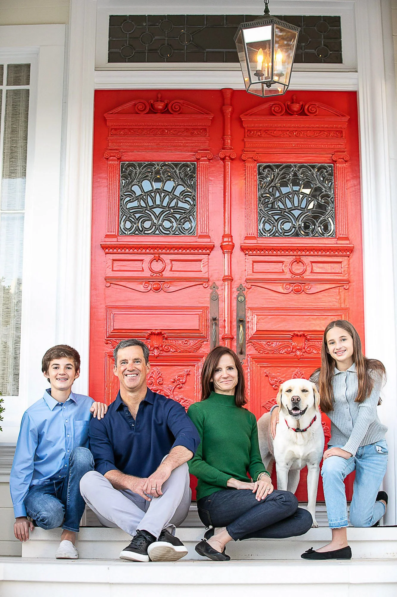 A family of five poses on the front porch of their house in front of a large red door with ornate black ironwork and decorative trim. The family includes a young boy, a middle-aged man, a woman, a teenage girl, and a Labrador retriever.