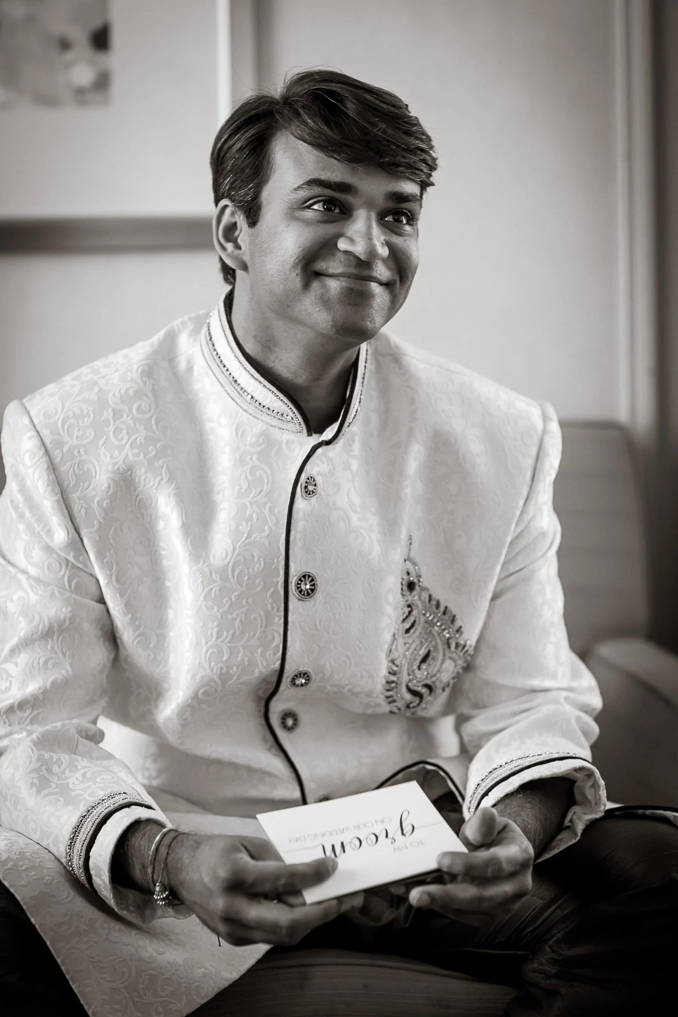 A young man with dark hair, wearing a traditional white embroidered shirt, sitting at a table, holding a card with the word 'greetings' in cursive, in a black-and-white photo.
