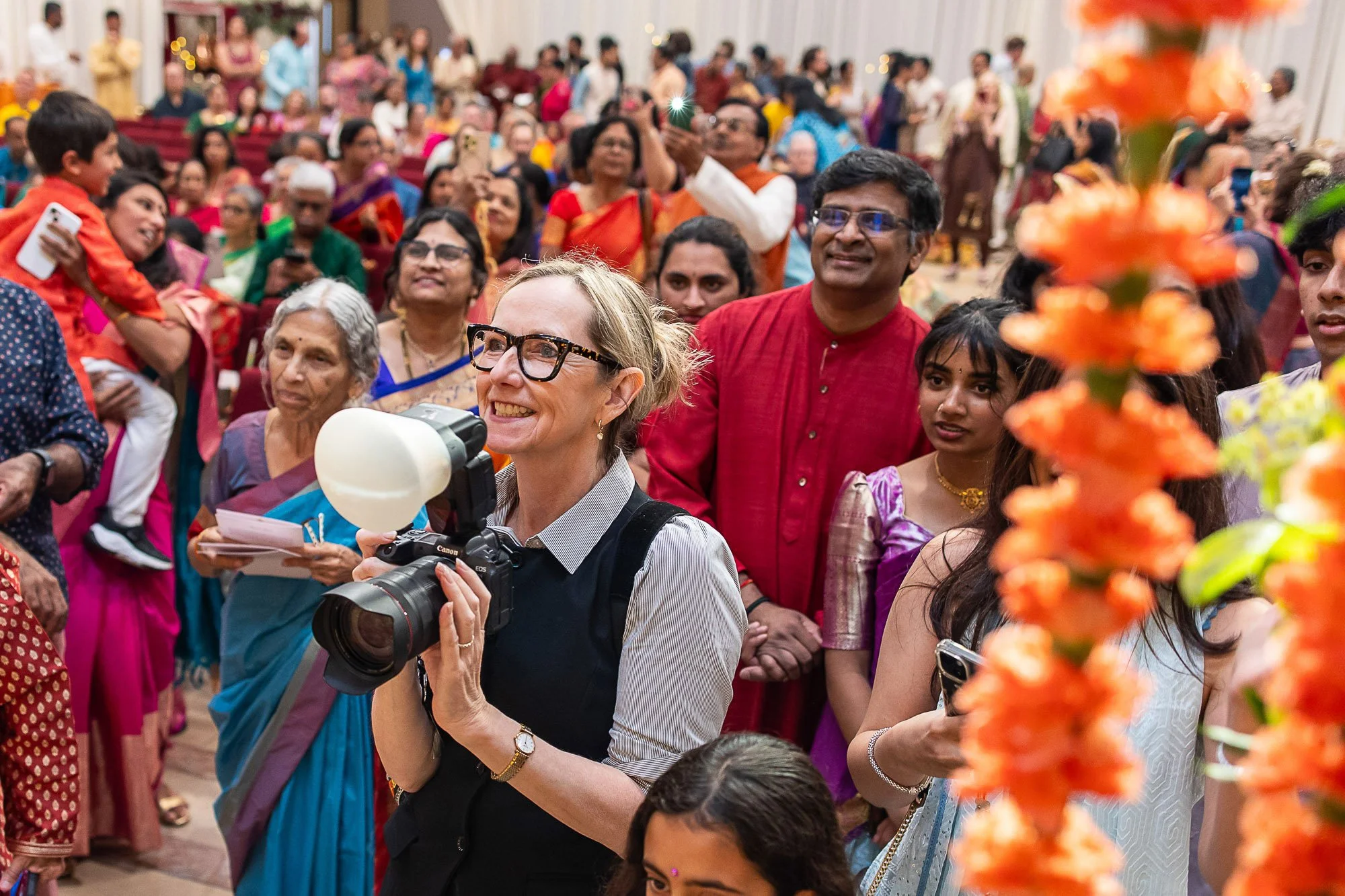 A diverse group of people, including women and children, attending an indoor cultural or festive event.  Tara Arrowood is holding a Canon camera with a flash attached, smiling and wearing glasses. 