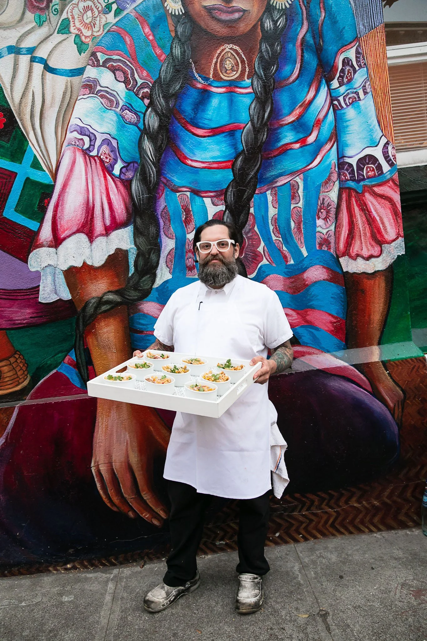 A man with tattoos, a beard, white glasses, and a white chef's coat holding a tray of small plated dishes, standing in front of a colorful mural painting that depicts a woman with long braided black hair, wearing a blue and red dress with floral and 