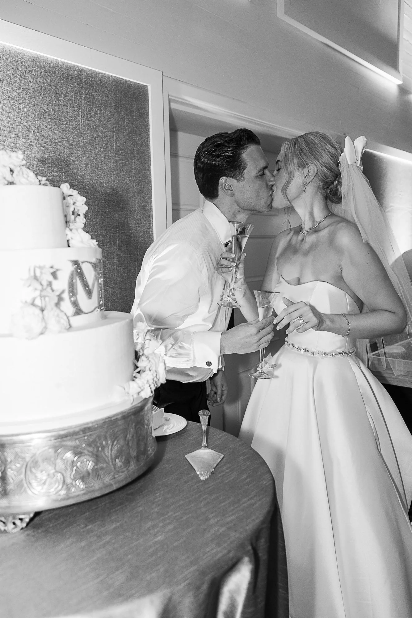 Black and white photo of a bride and groom sharing a kiss at a wedding reception, holding champagne glasses. The groom is dressed in a white shirt, and the bride is in a strapless wedding dress with a veil and jewelry. A wedding cake is partially vis
