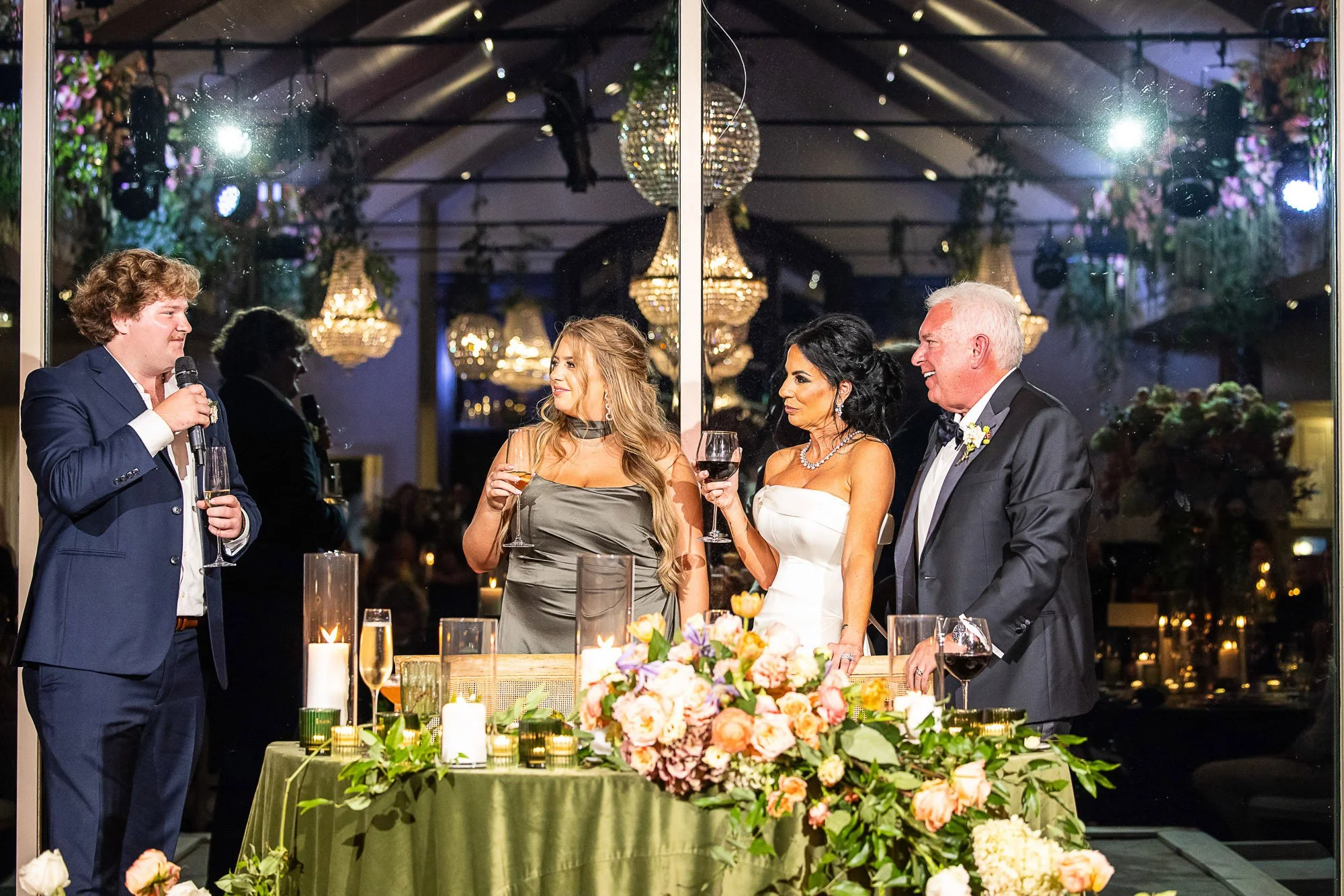 People standing around a decorated table at an elegant wedding reception, with chandeliers hanging from the ceiling and flowers in the foreground at the Beach and Tennis Club in Pebble Beach, CA.